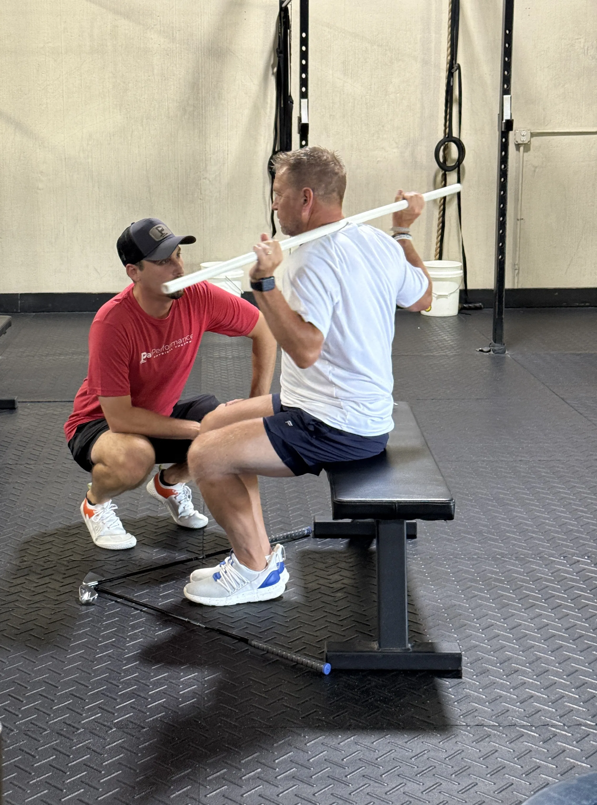 Man in white shirt sitting on a bench holding a long white stick behind his shoulders while another man in a red shirt and cap kneels in front, appearing to coach or assist in a gym.