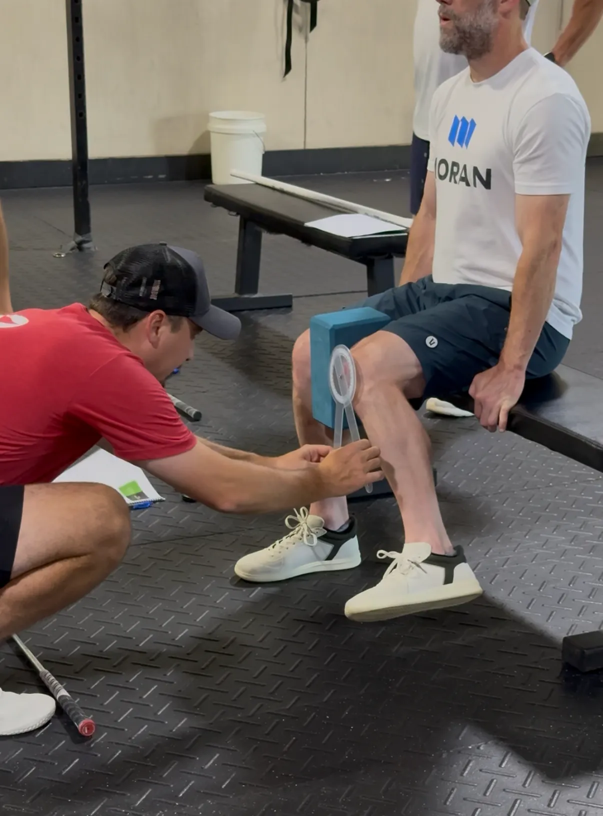 A man in a red shirt and black cap measures knee angle of a seated man with a goniometer in a gym setting.