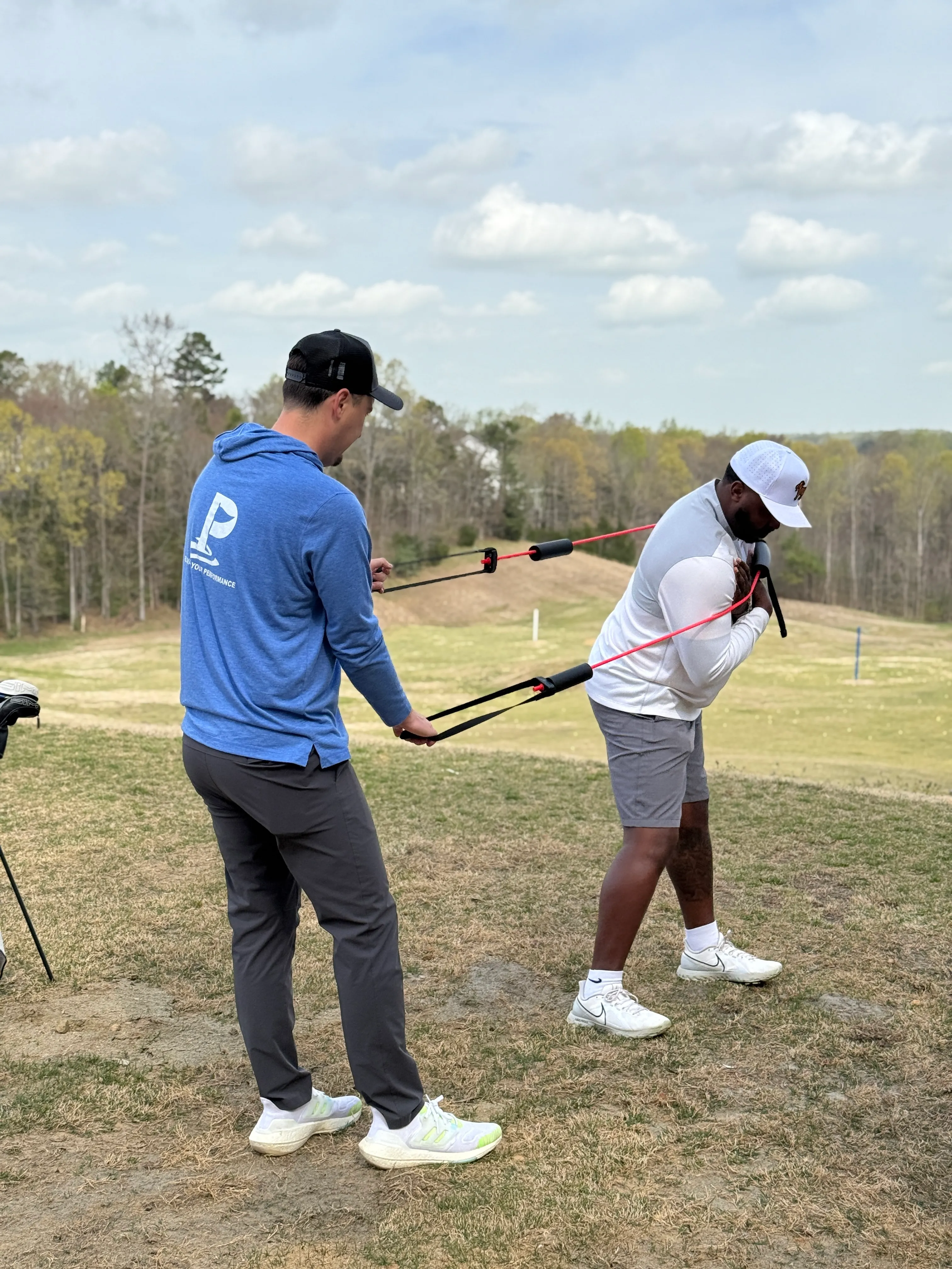 Two men outdoors on grass using resistance bands for golf swing training, one pulling while the other holds the bands.