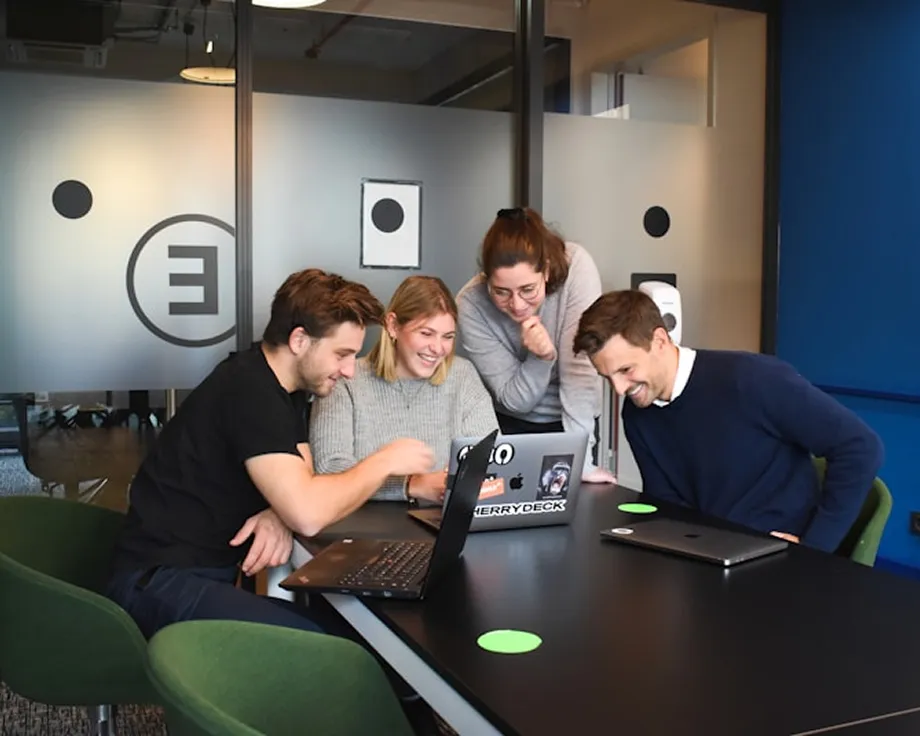 Four young professionals smiling and working together on laptops at a conference room table.