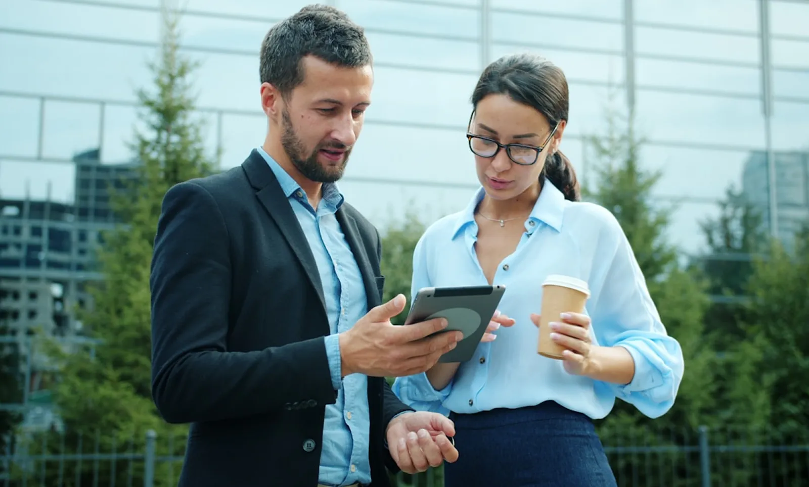 Man and woman standing outdoors looking at a tablet, woman holding a coffee cup.