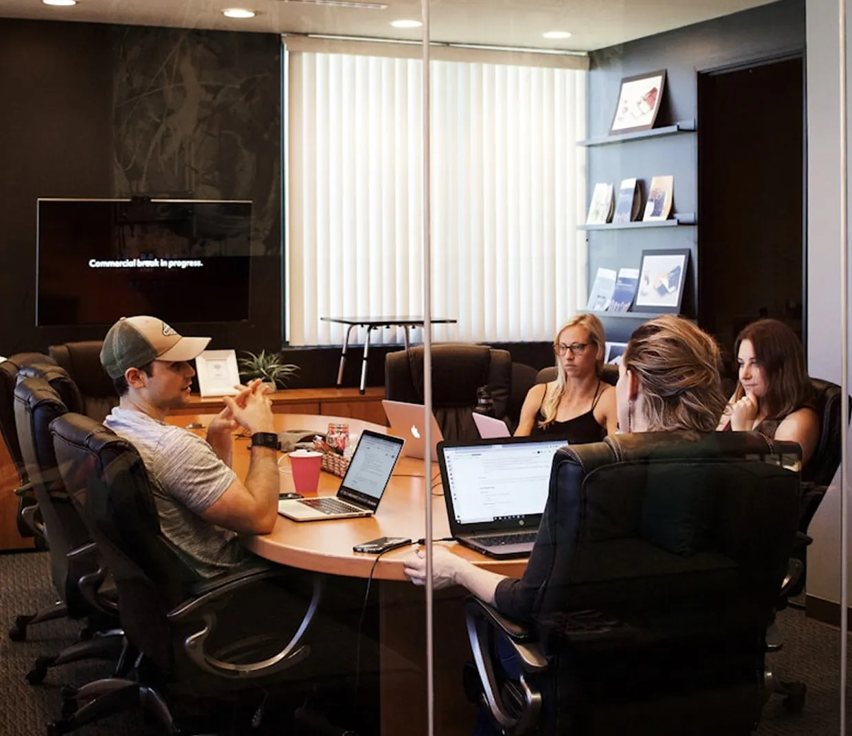 Four people in a conference room meeting around a table with laptops, a TV shows a commercial break.