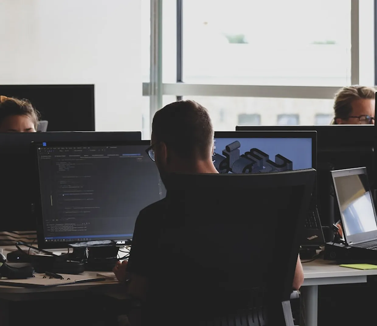 People working at desks with multiple computer monitors in an office with large windows.