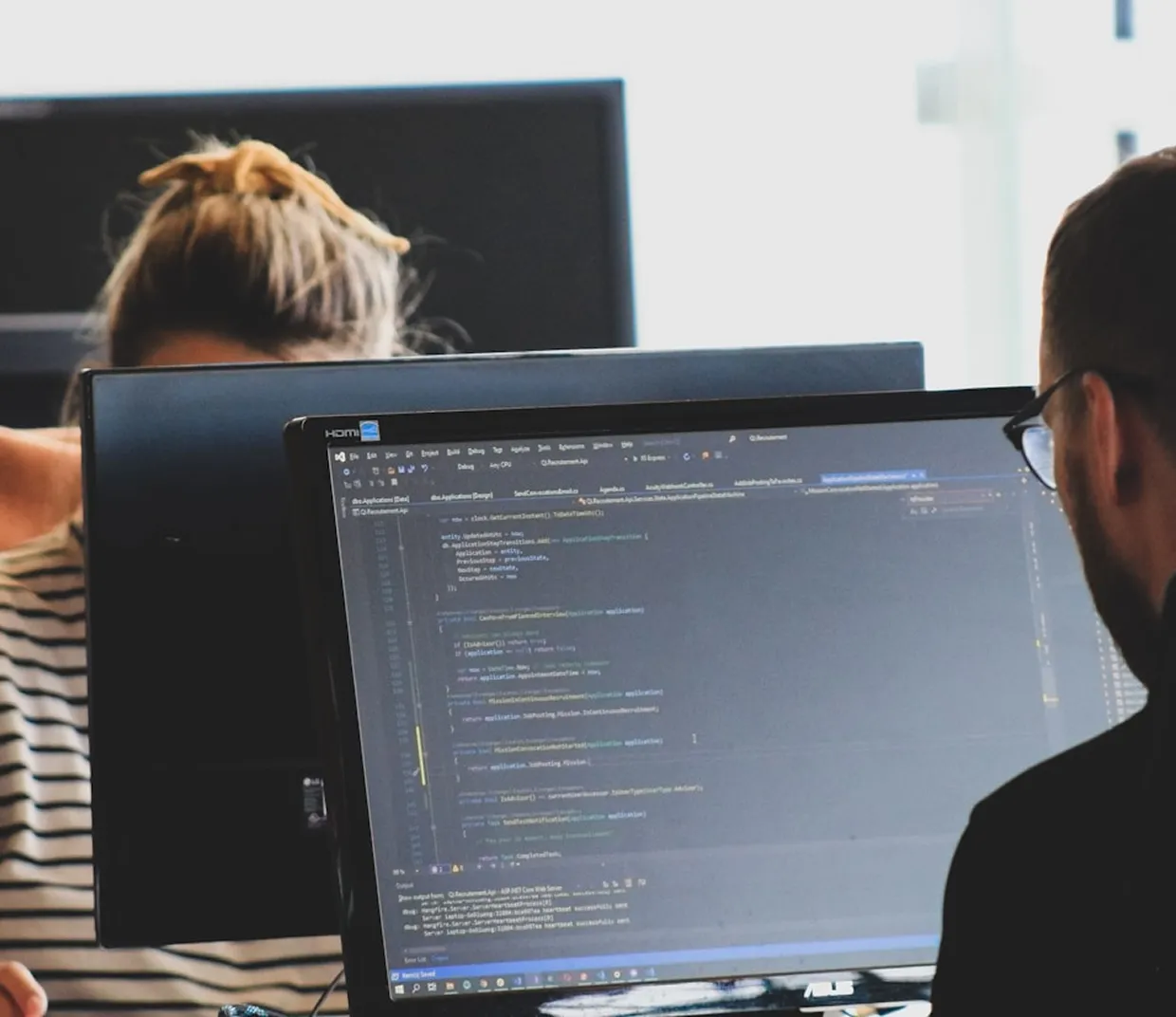 Man and woman working on computers, man viewing code on a monitor in an office setting.