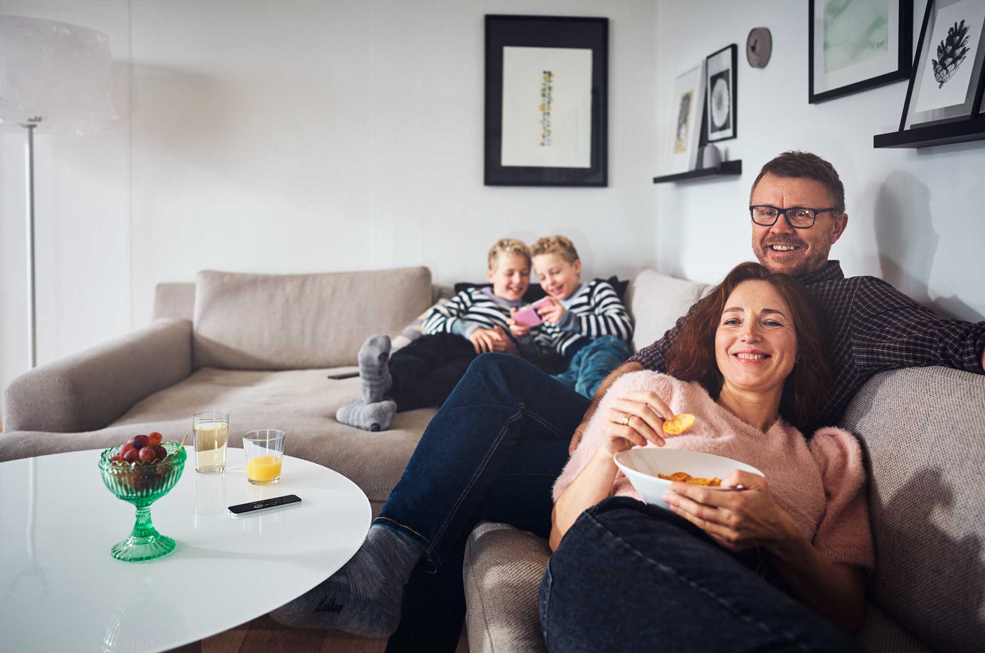 Familie koser seg i stuen med snacks og drikke, to barn ser på en mobiltelefon.