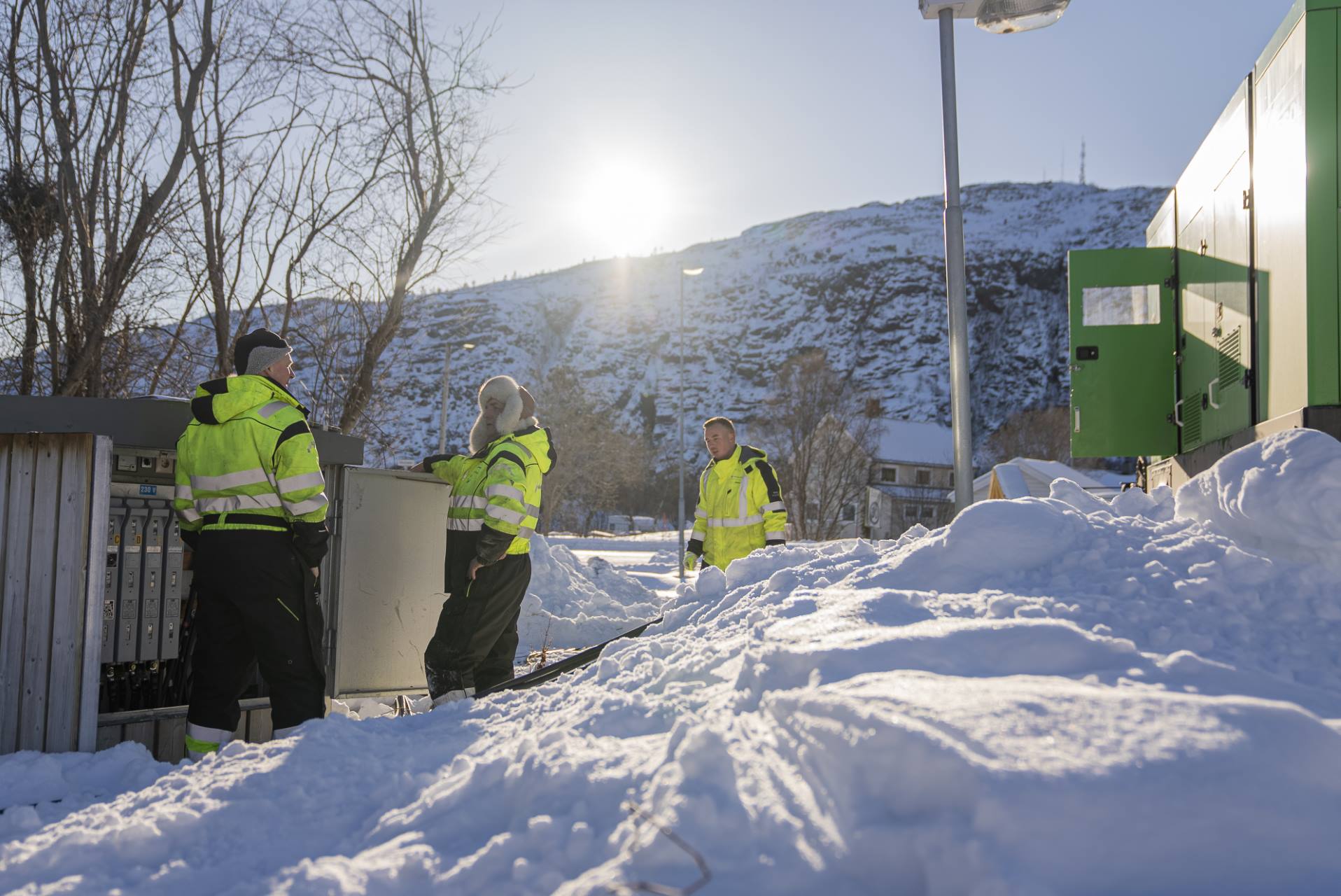 Tre arbeidere i refleksjakker jobber utendørs i snø med elektrisk utstyr under solen.