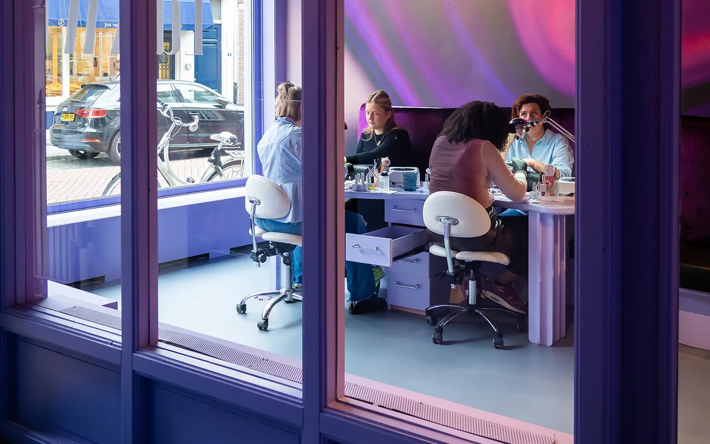 Nagelstylisten behandelen klanten in nagelstudio Lakwerk, gezien door het etalageraam van de salon