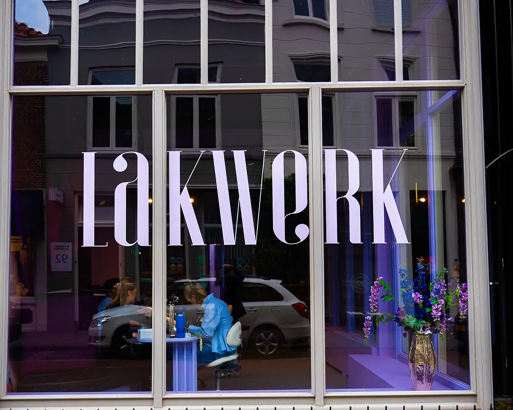 Buitenkant van nagelstudio Lakwerk in Amsterdam met het logo op de etalageruit en nagelstylisten zichtbaar aan het werk