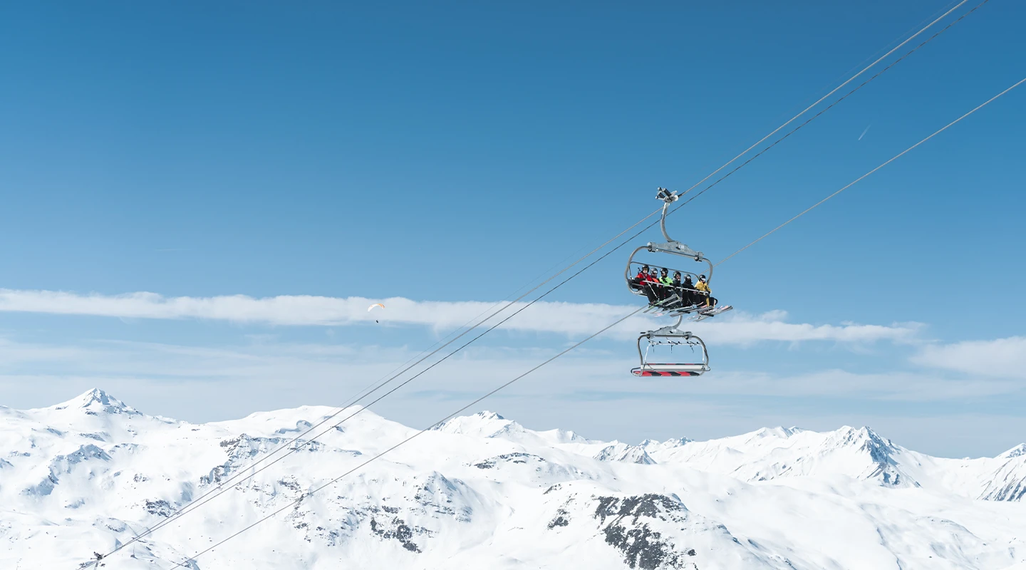 Skieurs sur un télésiège du domaine des Menuires avec en arrière plan les montagnes du massif et un parapentiste