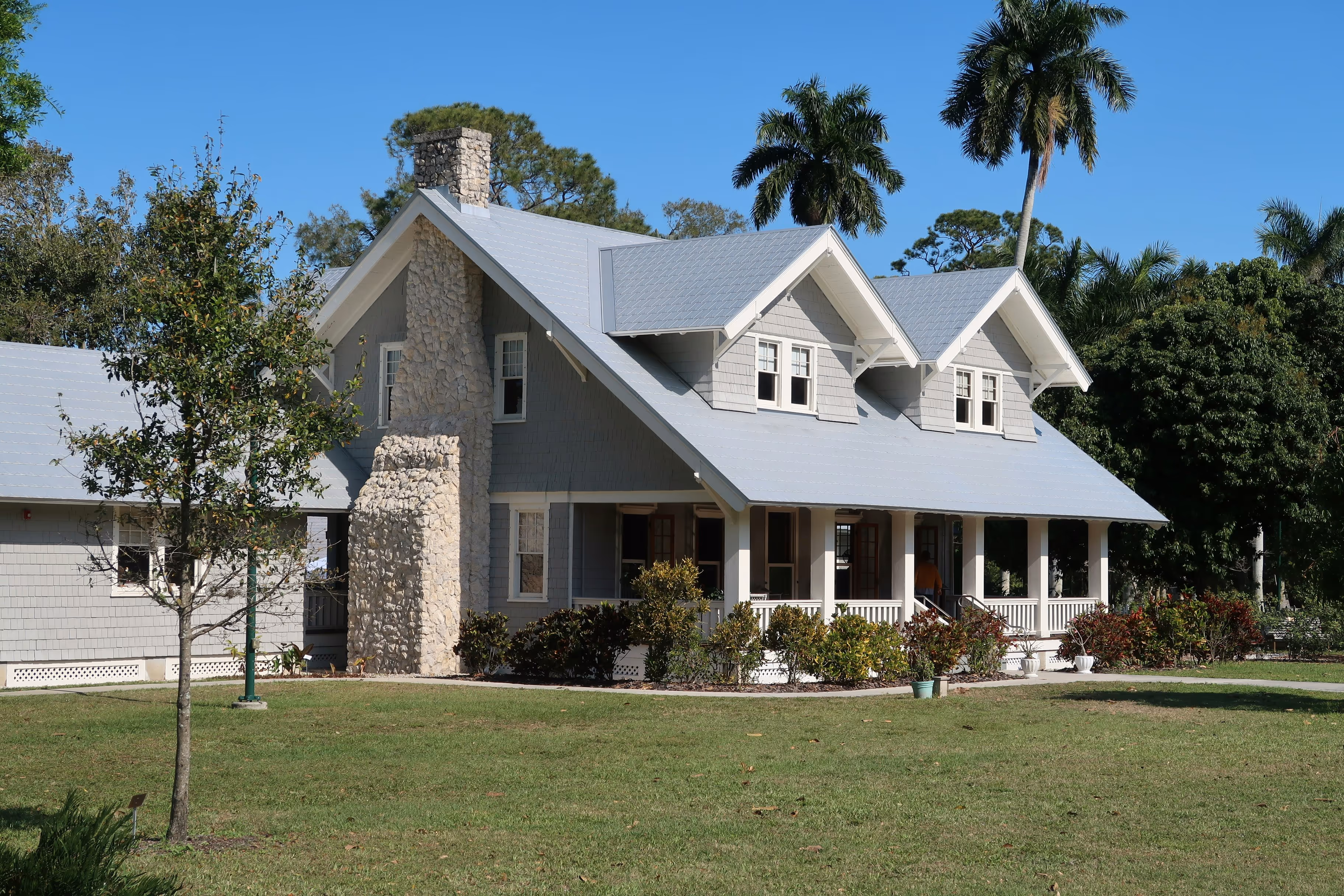 Gray craftsman-style home with a stone chimney, covered wraparound porch, and dormer windows, landscaped with palm trees