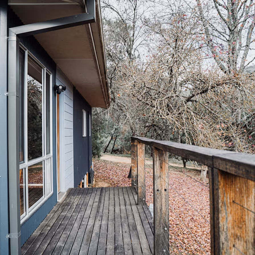 Covered wooden deck along the side of a home with large windows, overlooking a wooded, leaf-covered yard in late autumn