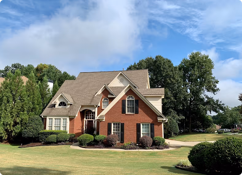 Two-story brick home with dark shutters, dormer windows, and a landscaped front yard bordered by mature trees