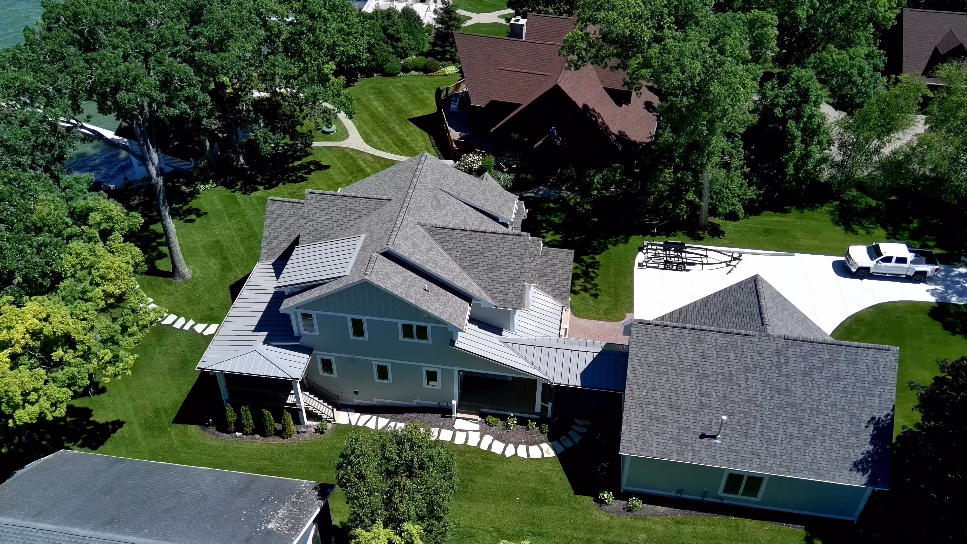 Aerial view of a large custom home with teal-blue siding and gray shingle roof, wraparound porches, and attached garage, surrounded by mature trees and lawn near a lake