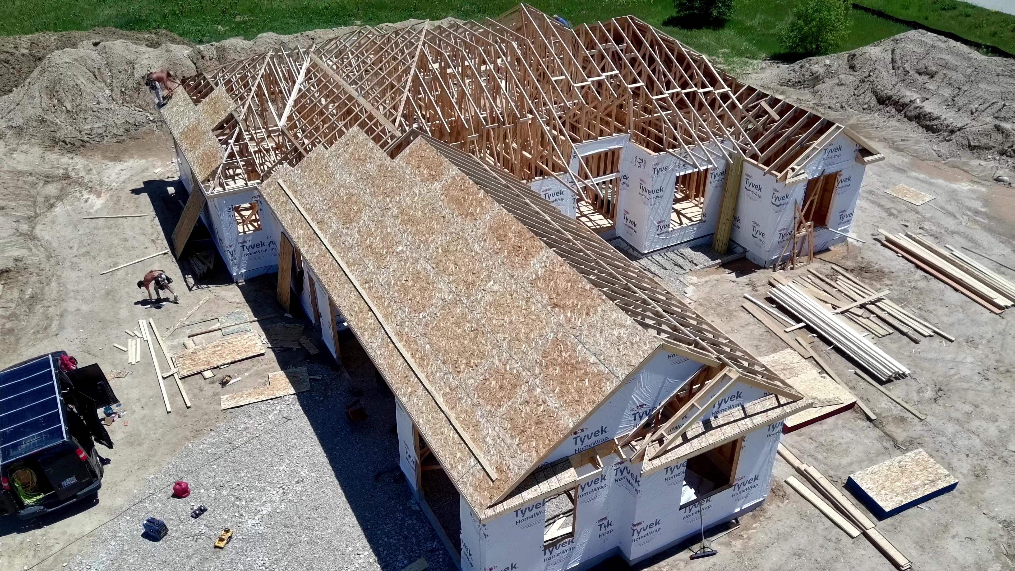 Aerial view of a custom home under construction with exposed roof trusses and Tyvek-wrapped exterior walls, building materials staged on the lot