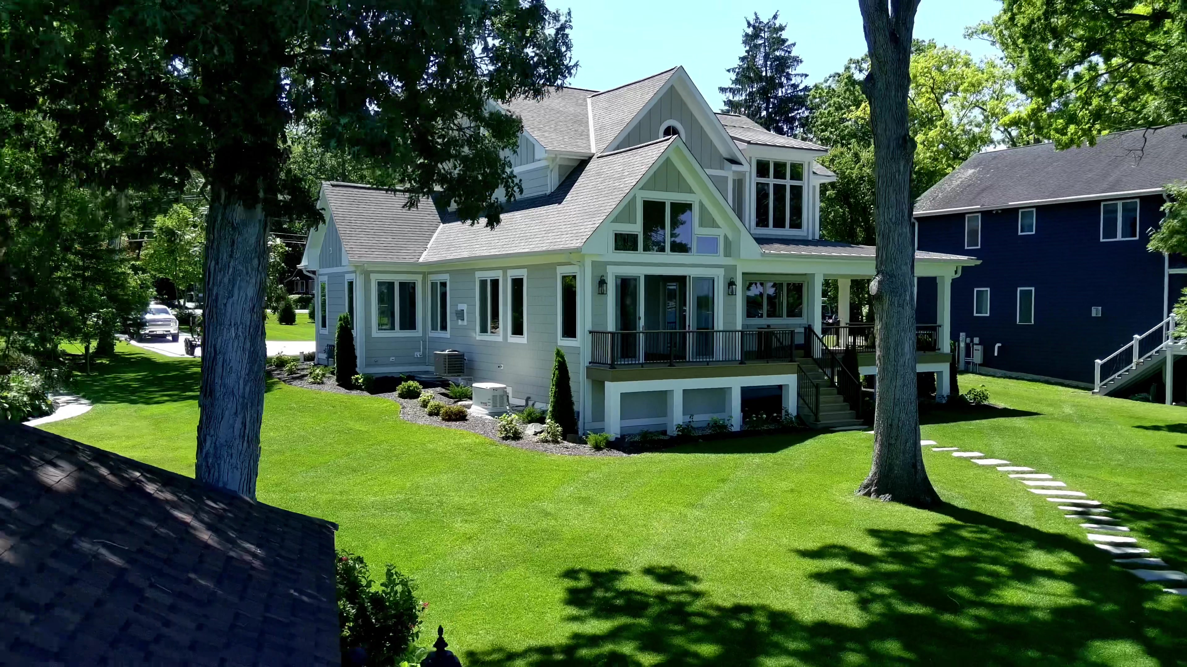 Aerial view of a finished custom lake home with light gray siding, covered porches, and a screened deck surrounded by green lawn and mature trees