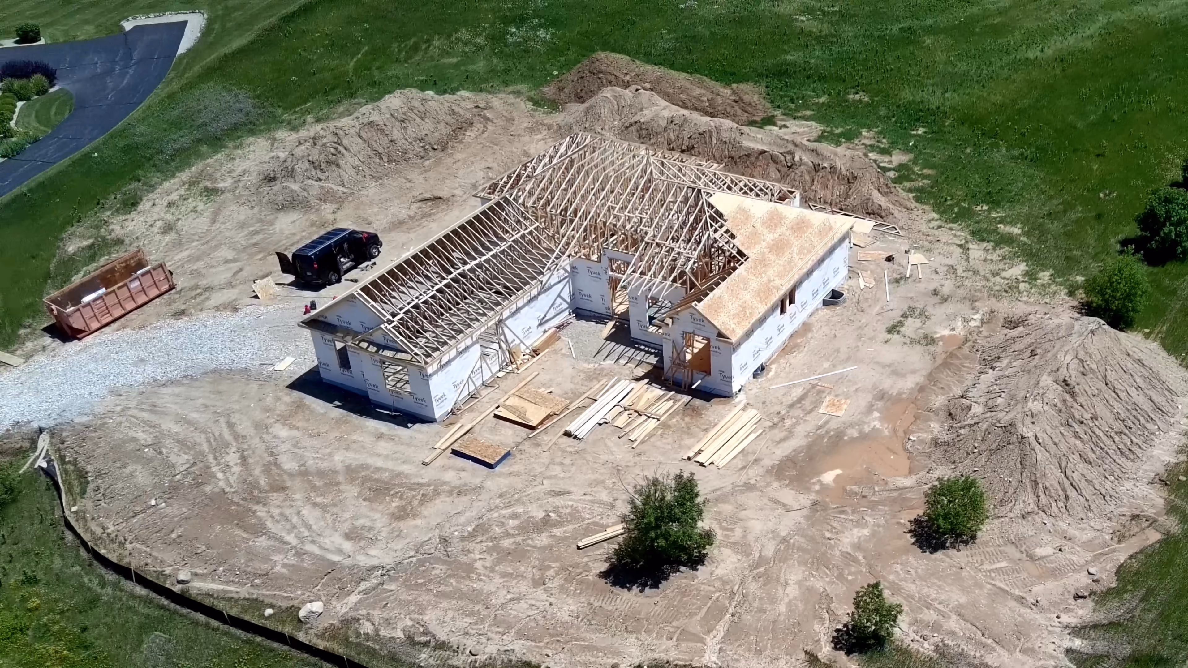 Aerial view of a custom home under construction with exposed roof framing, a gravel driveway, and a pickup truck on site