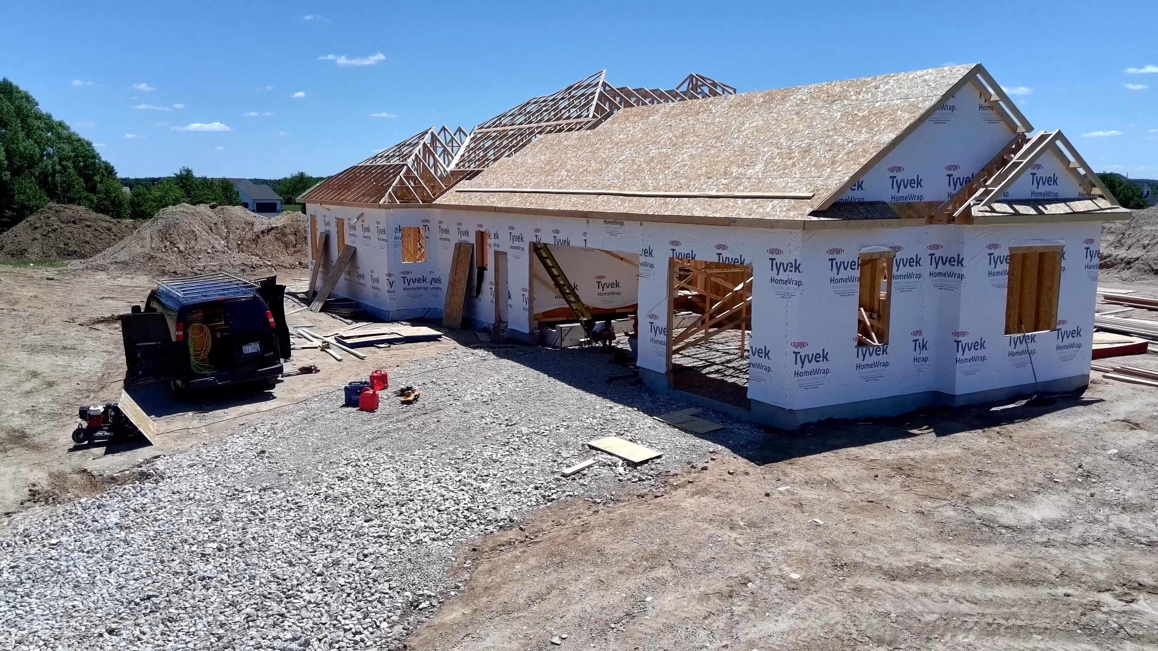 Aerial view of a custom home under construction with Tyvek-wrapped exterior walls, roof framing, and a vehicle parked on the gravel lot