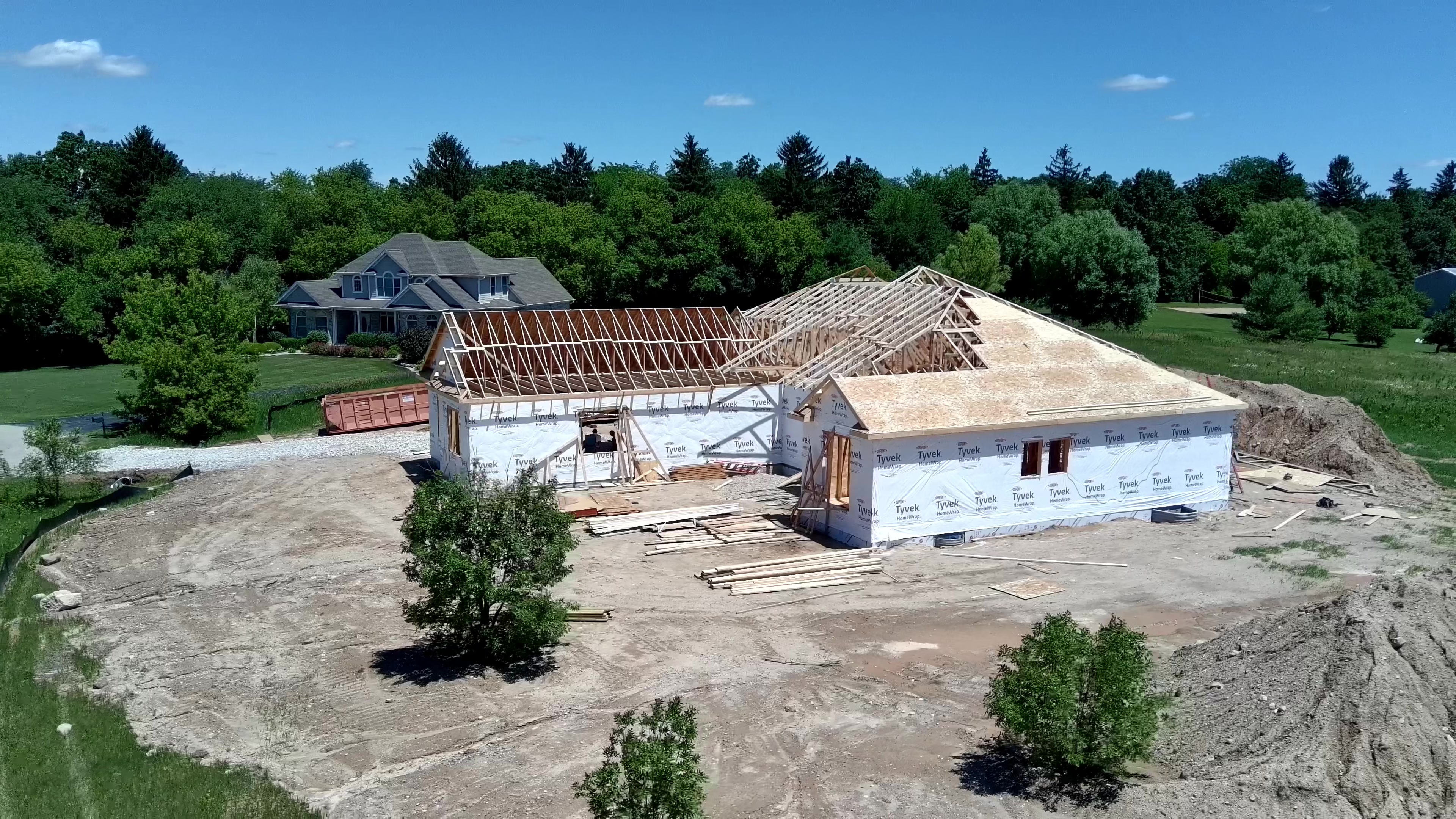Aerial view of a custom home under construction with framed roof trusses and wall sheathing, near a finished house on an open lot