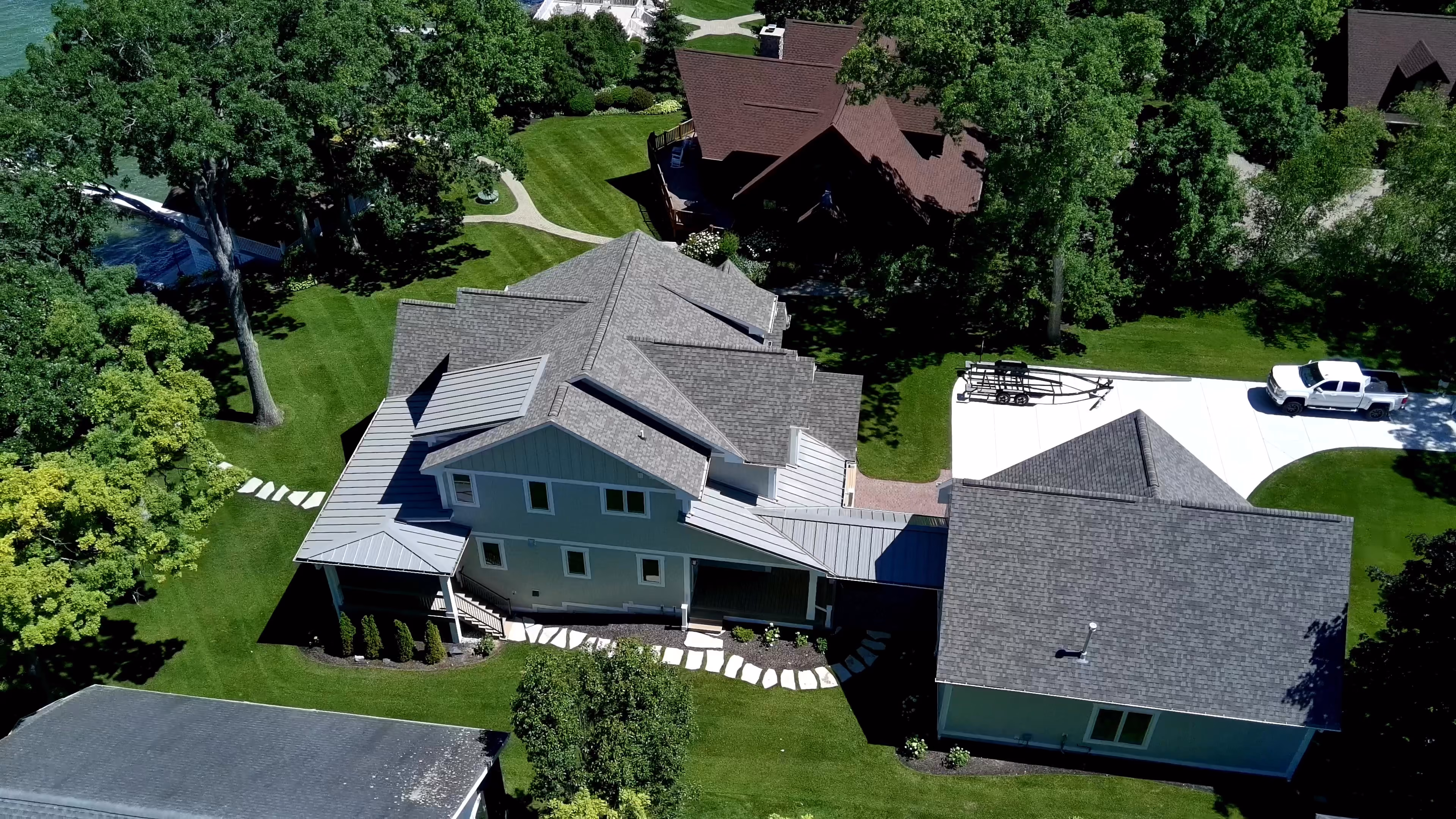 Aerial view of a large custom home with teal-blue siding and gray shingle roof, wraparound porches, and attached garage, surrounded by mature trees and lawn near a lake