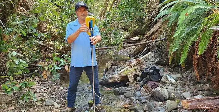 Worker holding surveying equipment beside a forest creek