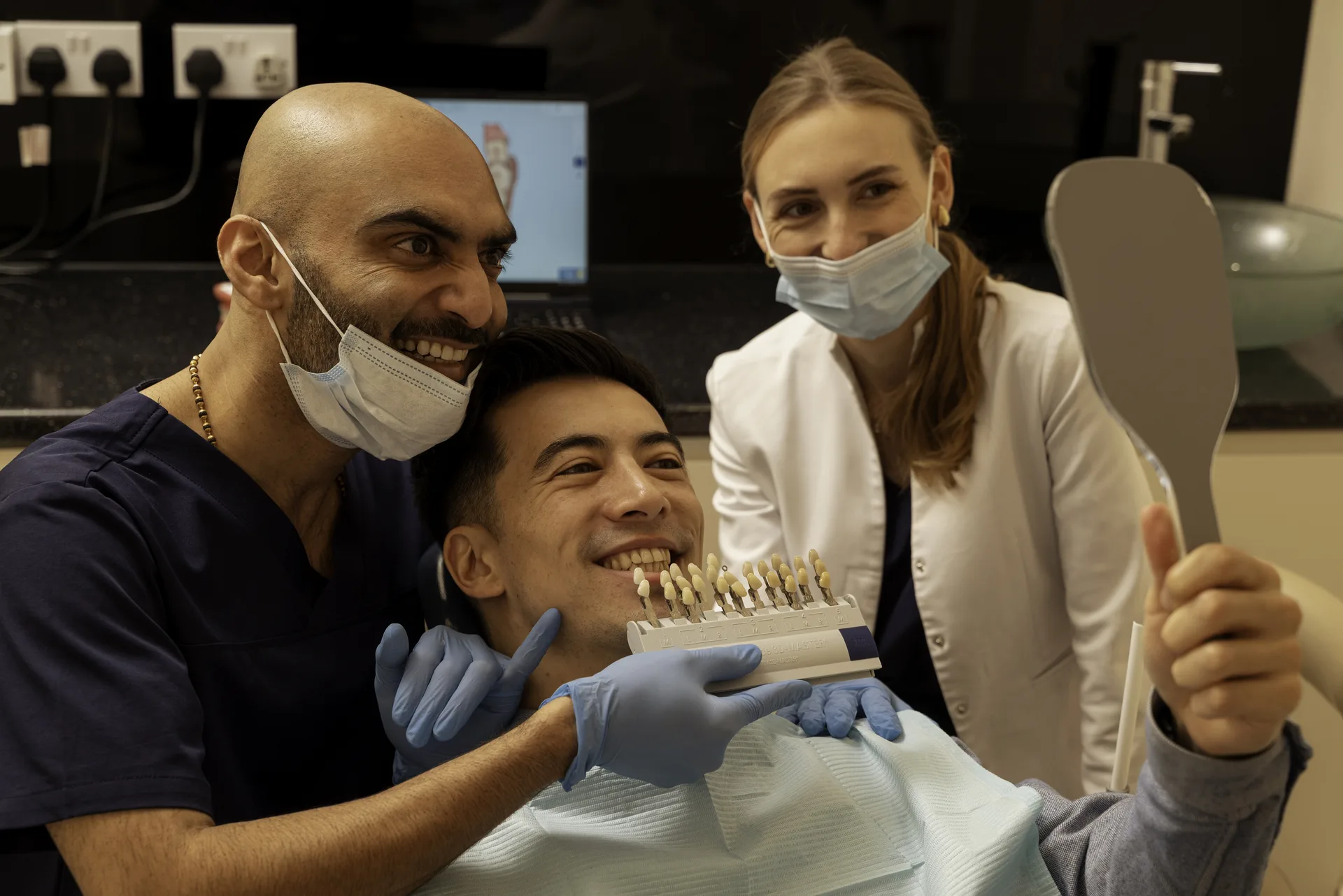 Teeth whitening cost: dentist and nurse with a smiling patient holding a mirror and shade guide