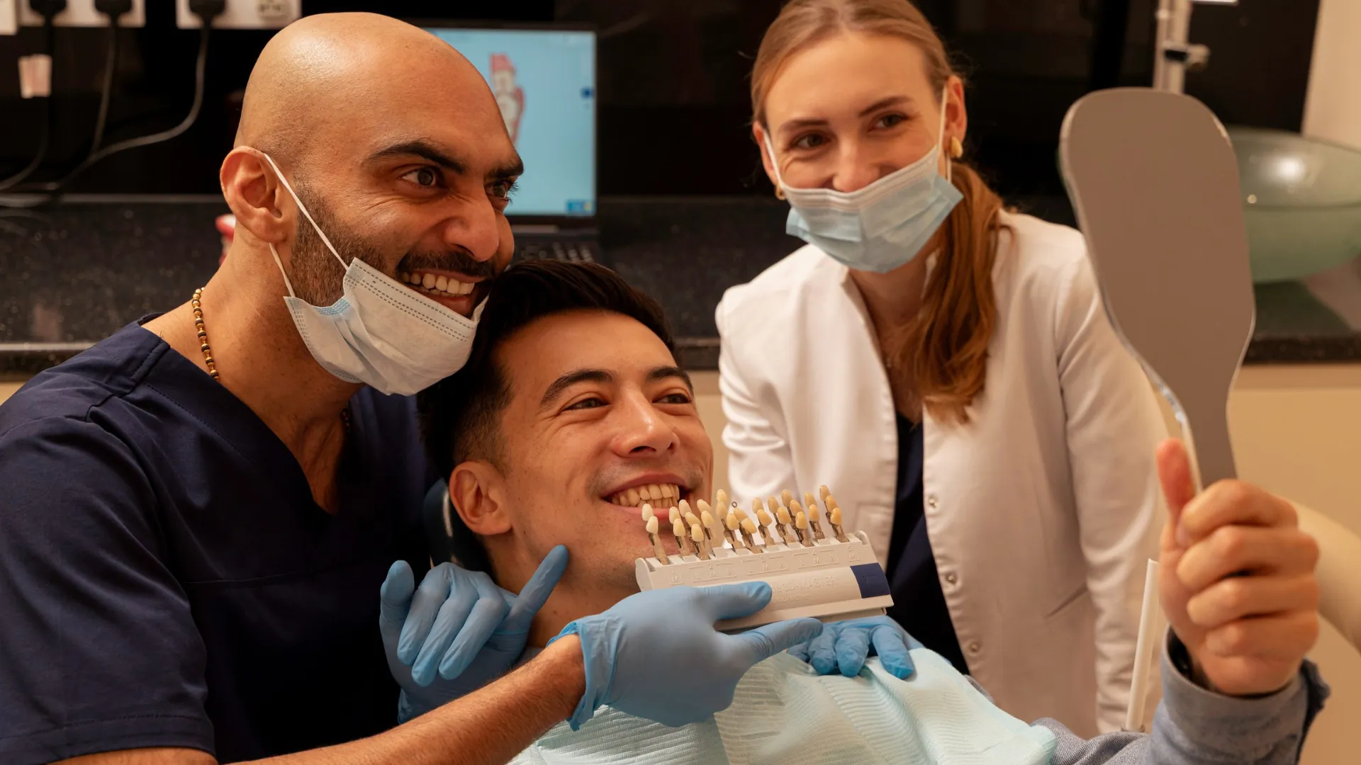 Discoloured teeth treatment: dentist and nurse with a smiling patient holding a mirror and shade guide after whitening