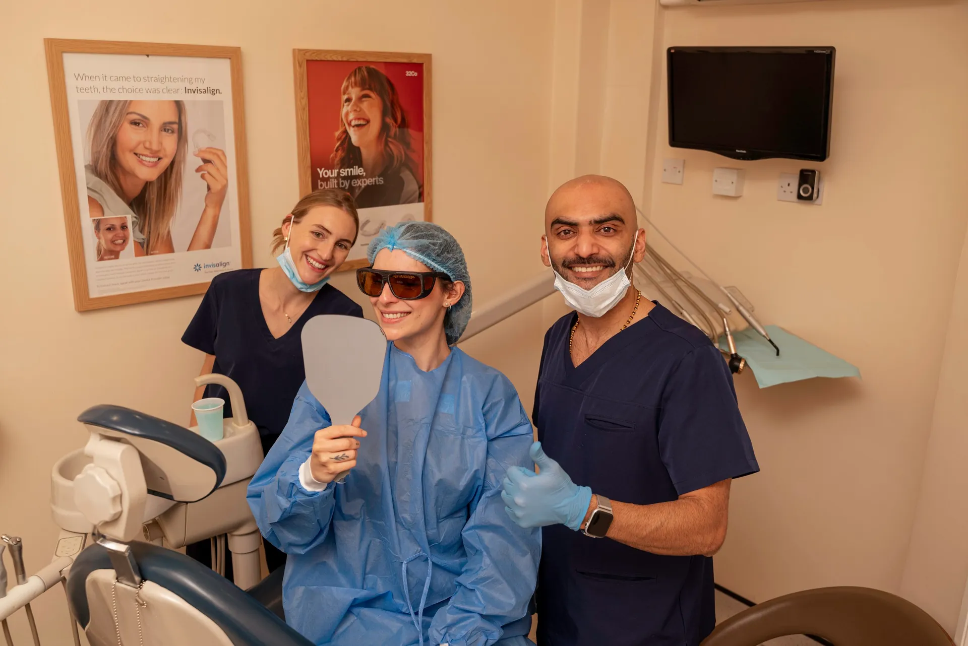 Dental team and a smiling patient in protective glasses holding a mirror after treatment — composite bonding longevity