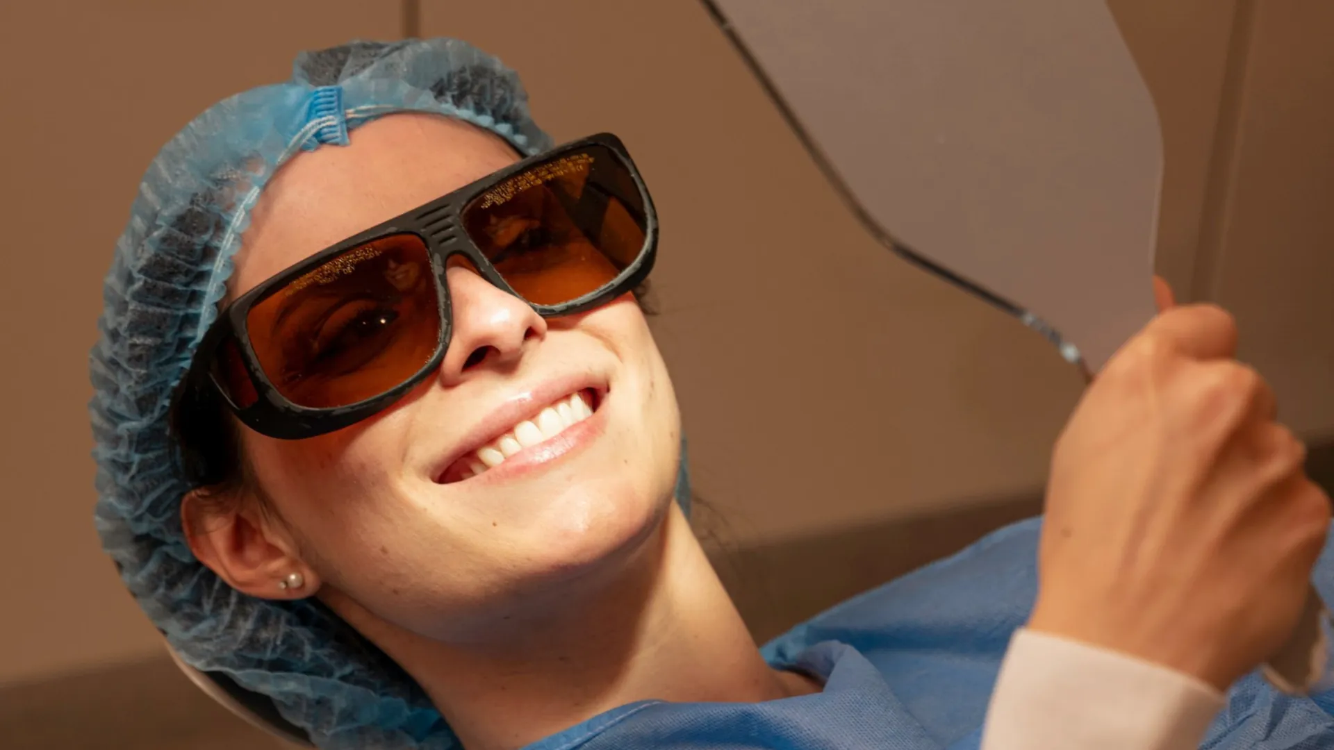 Smiling patient in protective glasses and a hair cap reclining for treatment