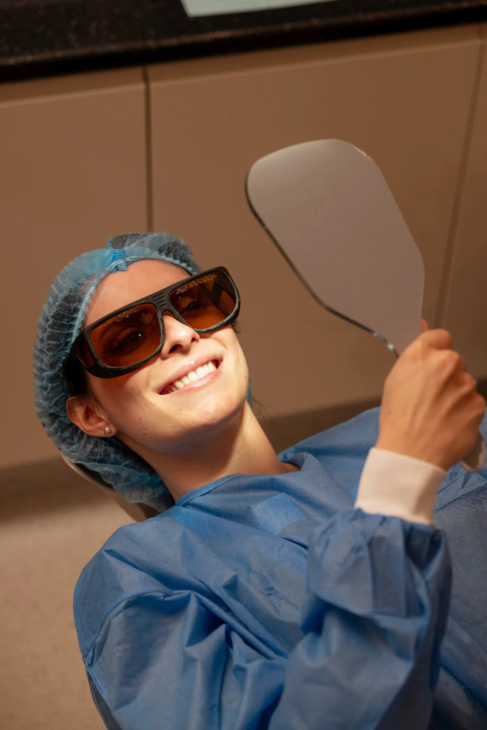 Smiling patient in protective glasses and a hair cap checking her teeth in a mirror — AHA BHA chemical exfoliation