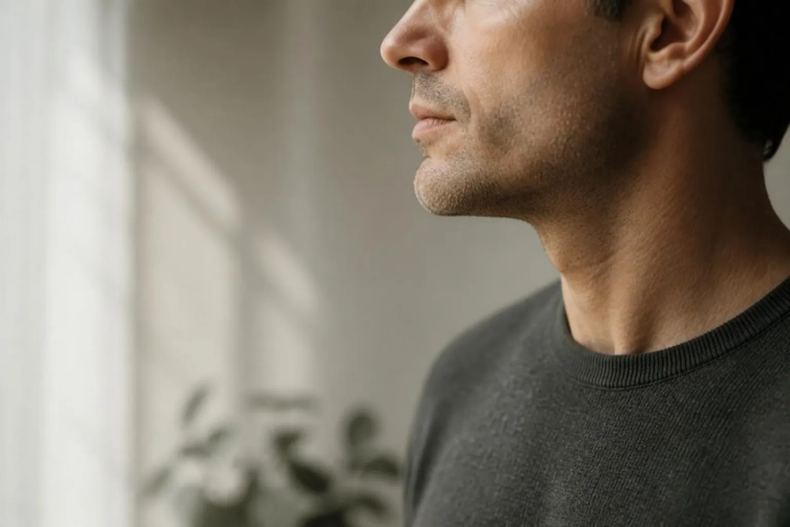 Side profile of a man's jawline and chin against a soft cream wall, natural light