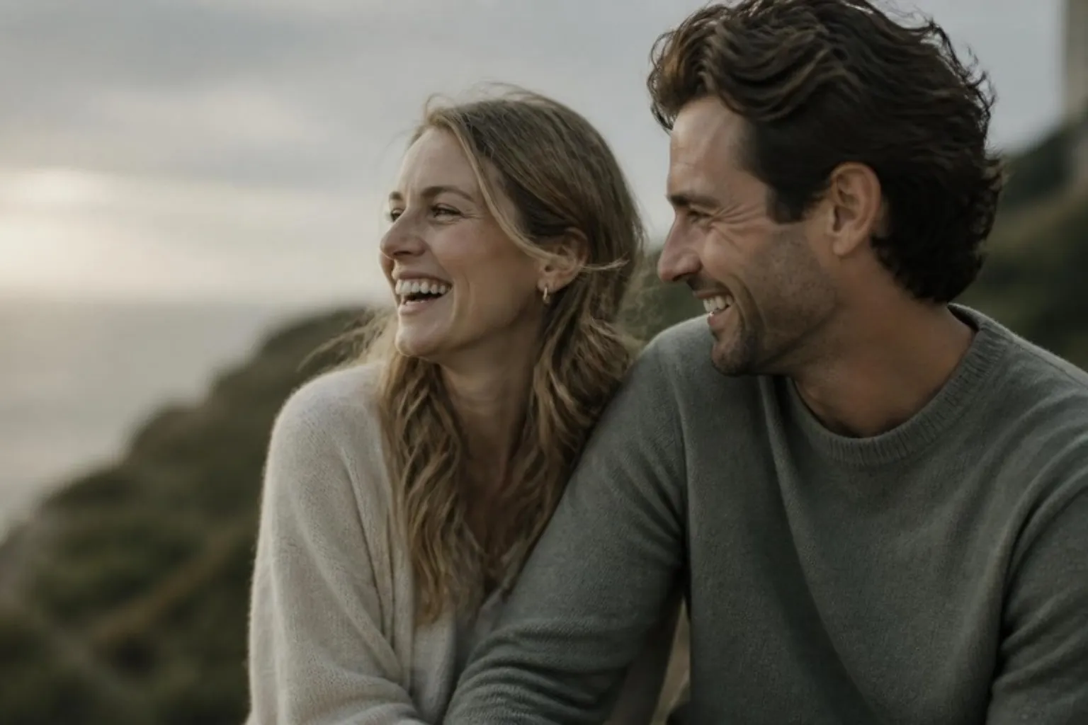 A couple laughing together outdoors in soft evening light, faces partly turned