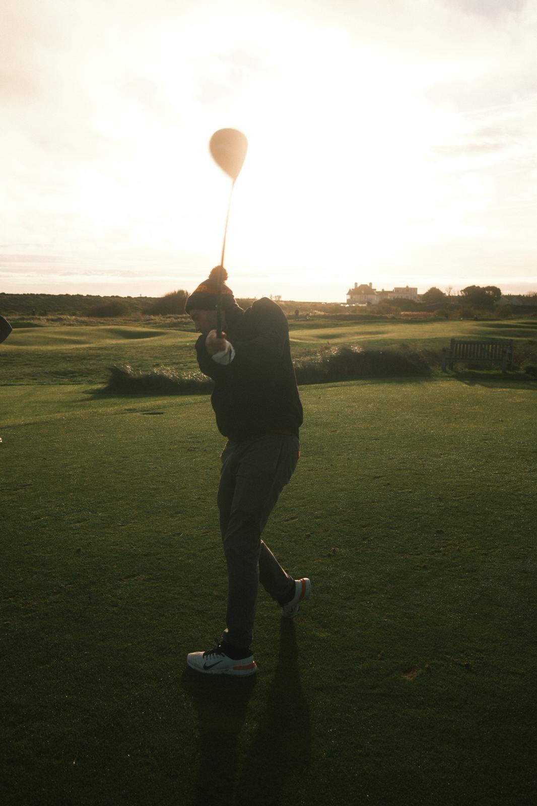 A golfer hitting a driver with a sunrise behind him on a links golf course