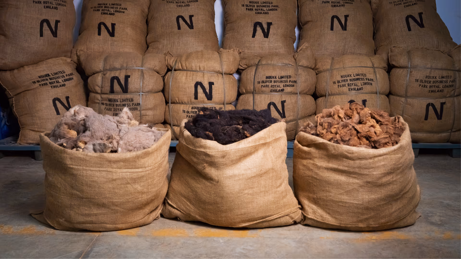 A warehouse wall of stacked NOUKK burlap sacks with three open bags in the foreground displaying raw Arabian camel hair in light, dark, and reddish-brown desert tones.