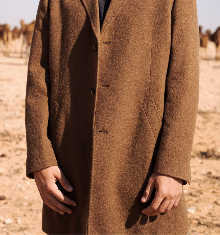 A model in a high-end NOUKK camel hair overcoat standing in a desert landscape with camels in the background, illustrating the heritage and premium drape of the 16-micron fiber.