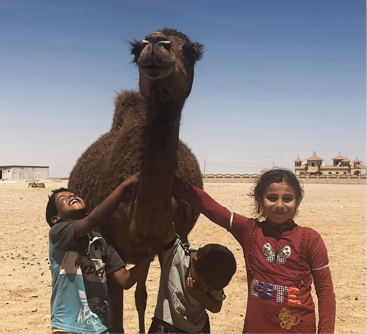 Nomadic children smiling and interacting with a camel in the desert, representing NOUKK's social impact and community empowerment initiatives.