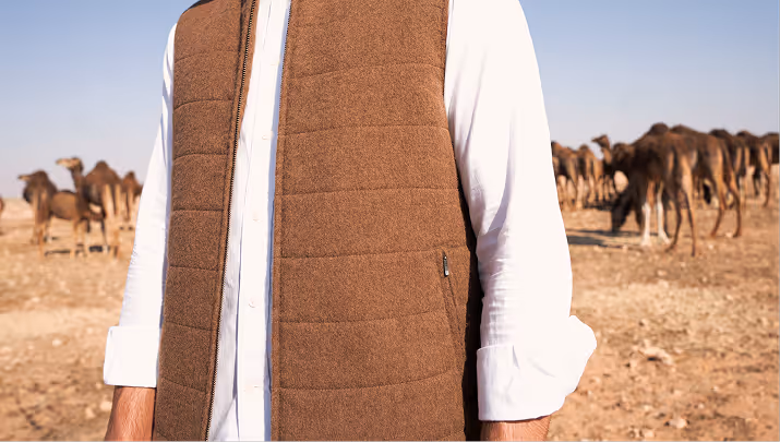 Model wearing a NOUKK camel hair garment in the Arabian desert with free-range camels in the background