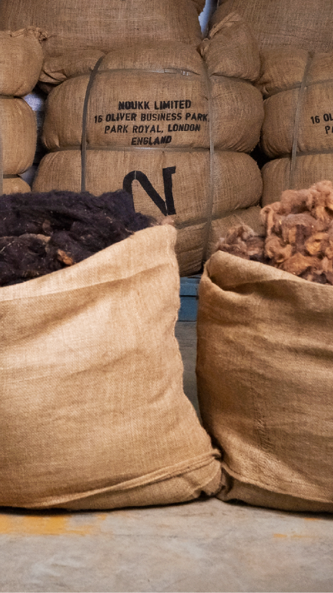 A warehouse wall of stacked NOUKK burlap sacks with three open bags in the foreground displaying raw Arabian camel hair in light, dark, and reddish-brown desert tones.