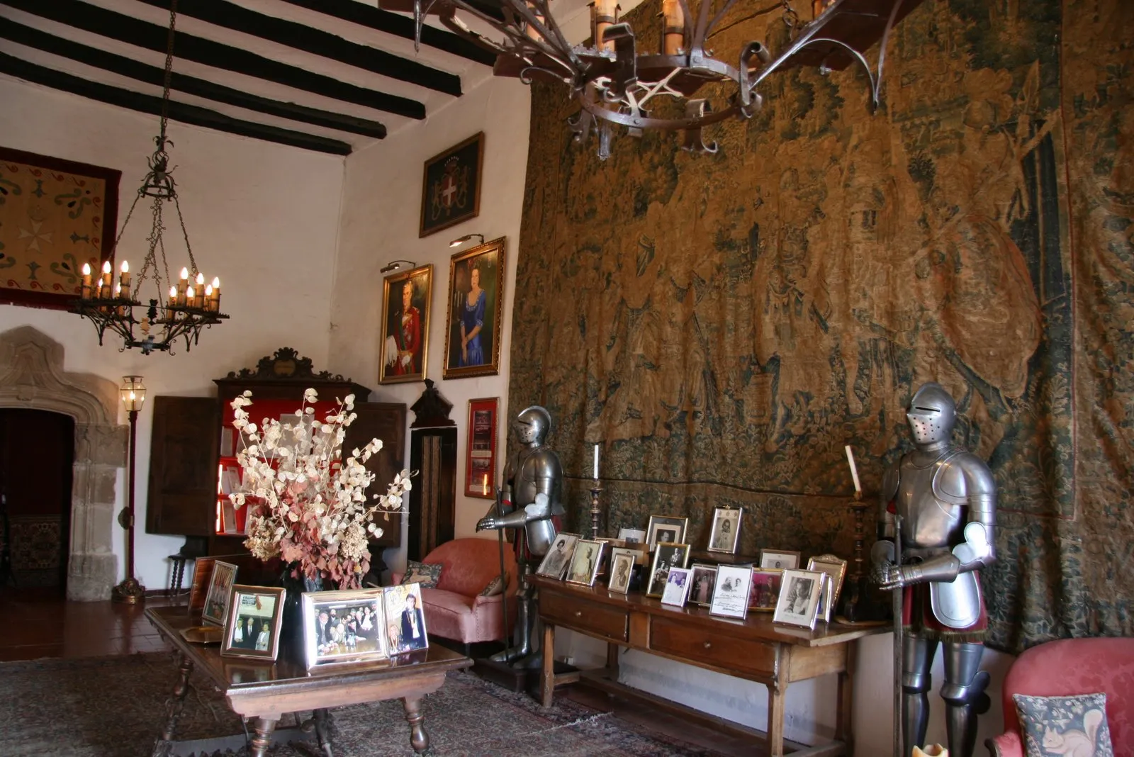 Sala d'estil antic amb armadura medieval de metall a banda i banda, una gran tapisseria a la paret i un taulell amb fotos enmarcades i un gerro de flors seques al centre.