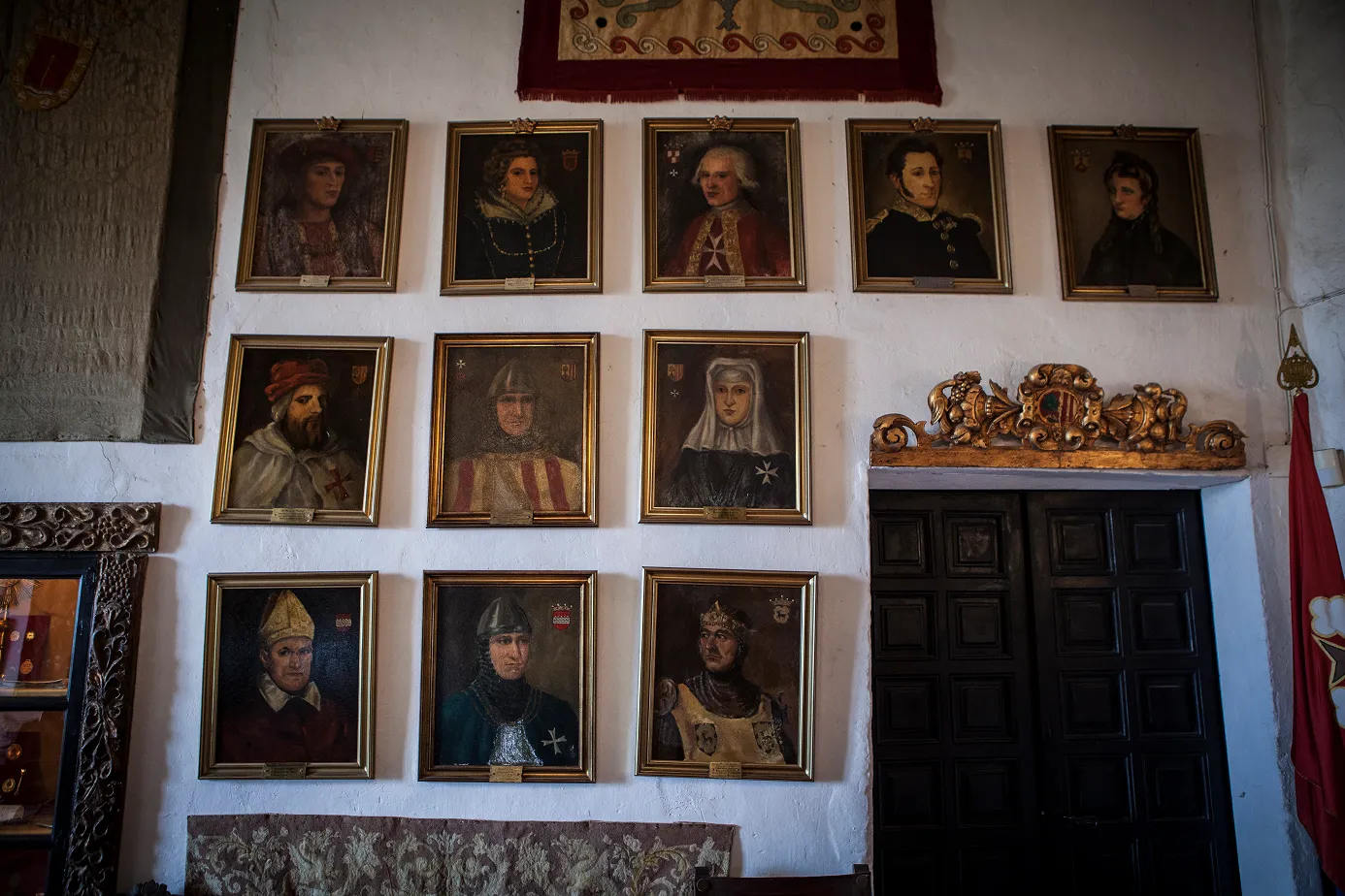 Una paret de paret blanca amb quinze retrats pintats en marc daurat de personatges històrics o nobiliars, la majoria amb vestits antics i motivos de cavallers, col·locats en tres files, al costat d'una porta de fusta fosca amb un ornament daurat a sobre.