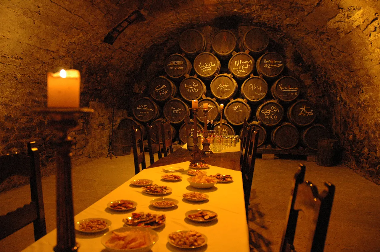 Sala amb taula llargada amb estovalles blanques, ple de plats amb aperitius, al centre espelmes en uns candelabres i a la paret de pedra de fons, una pila de barrils de vi signats, il·luminat amb llum càlida.