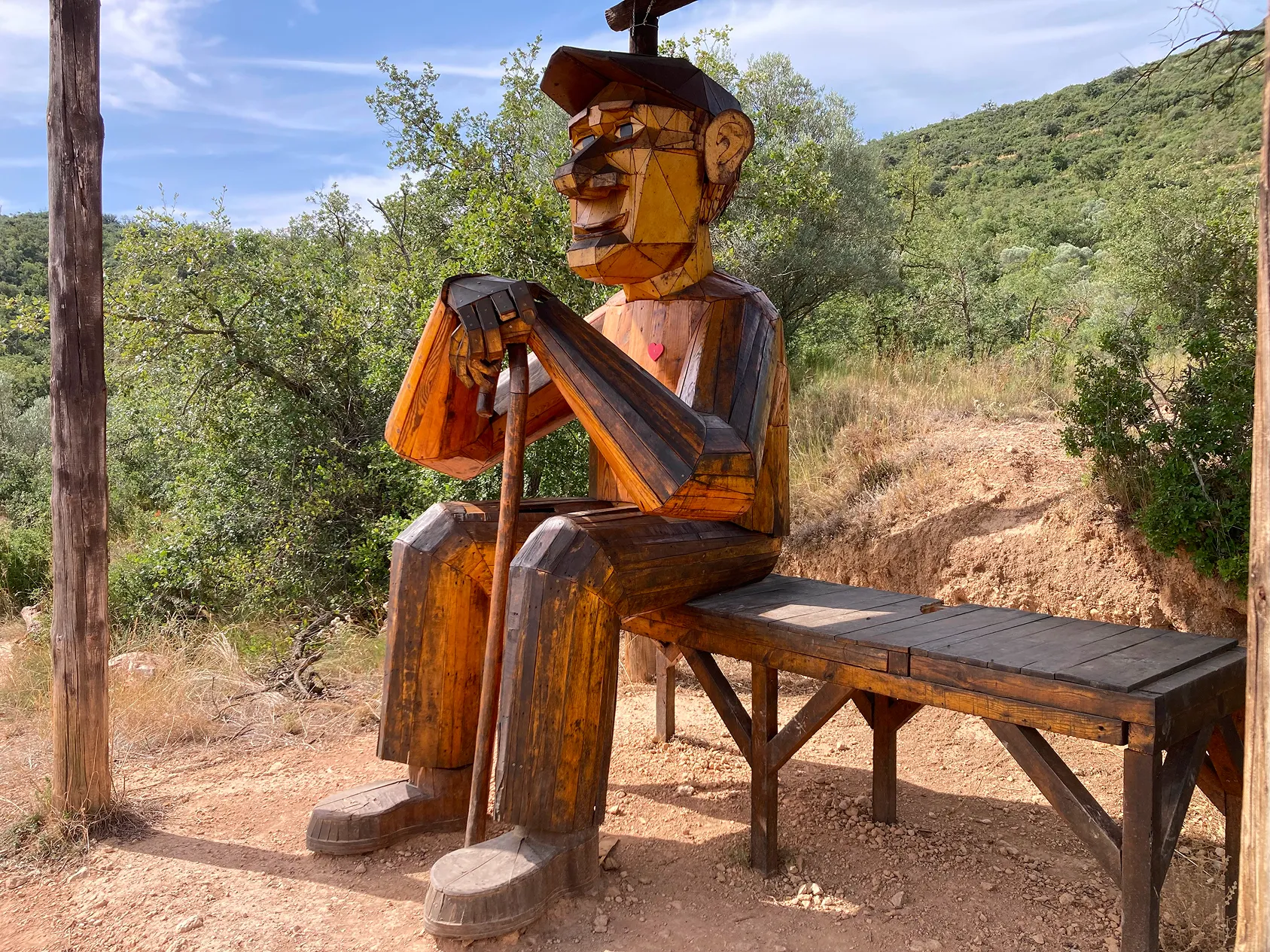Escultura de fusta geomètrica d'una figura humana asseguda amb un bastó sobre un banc de fusta en un entorn natural amb arbres i muntanyes.