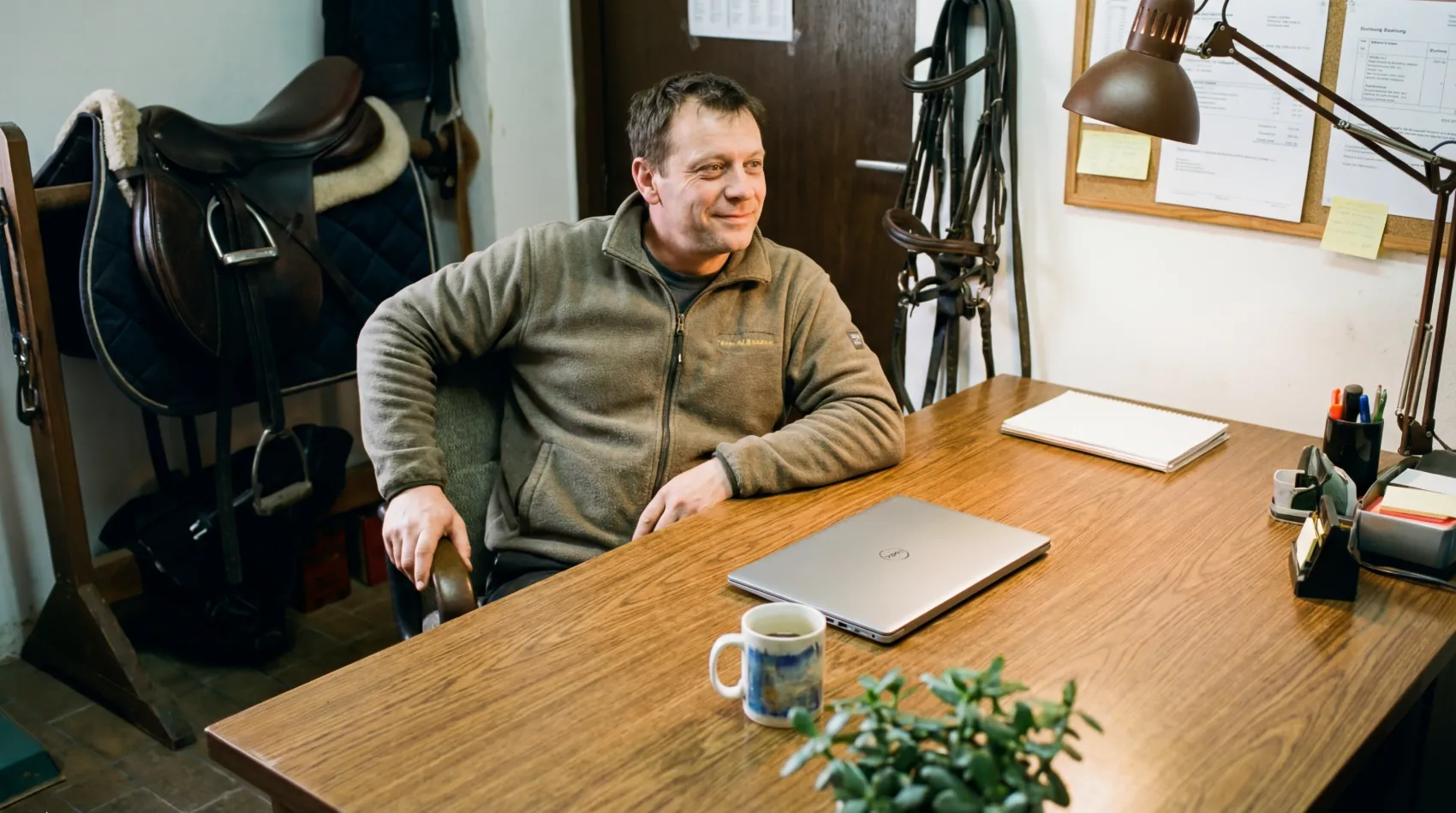 Entspannter Stallbetreiber am aufgeräumten Schreibtisch mit Laptop im Stallbüro — organisierter Arbeitsalltag dank digitaler Stall Software equitano