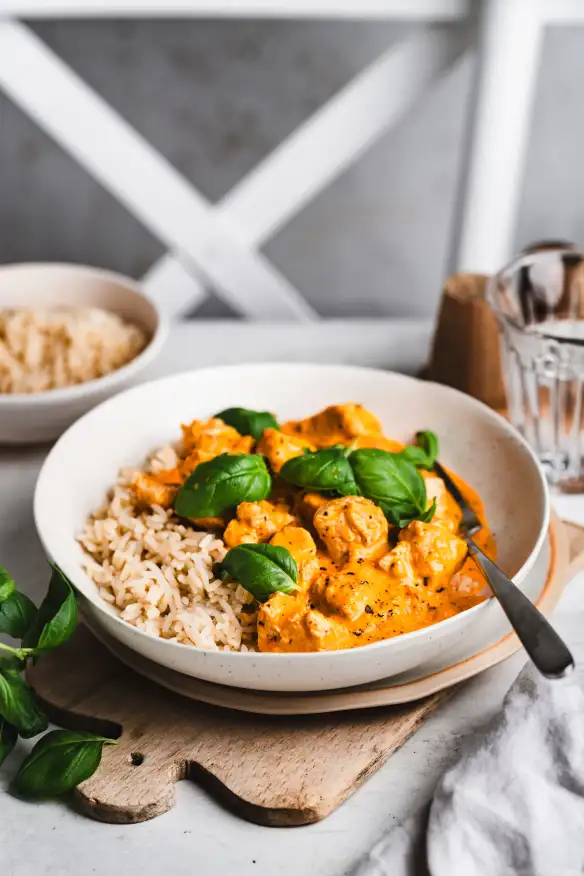 Bowl of rice topped with creamy orange chicken curry garnished with fresh basil leaves on a wooden board.