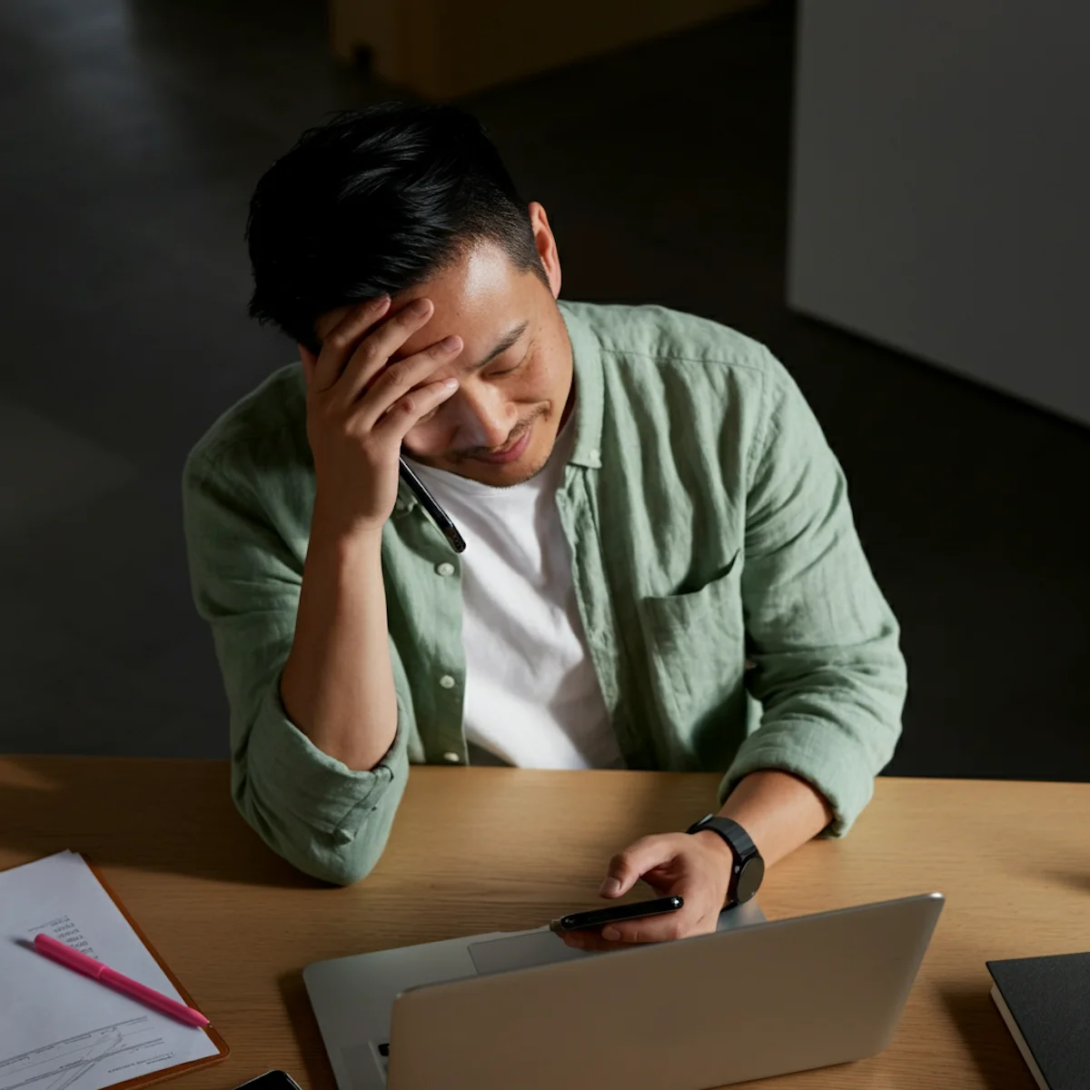 Mann hält Handy und Telefon am Ohr, wirkt gestresst, sitzt vor Laptop an Schreibtisch.