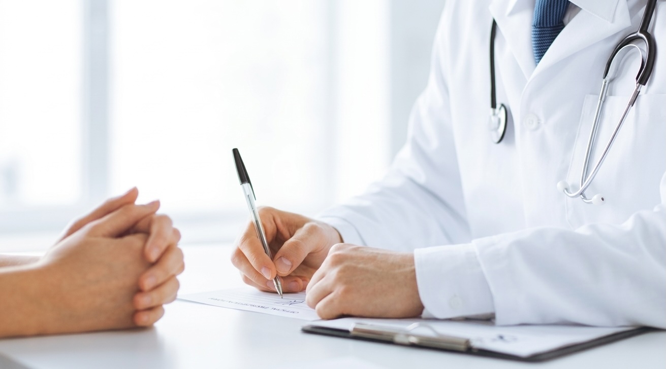 Doctor in white coat with stethoscope writing on a clipboard while patient with clasped hands listens.