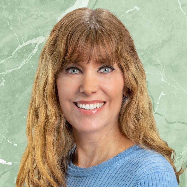 Smiling woman with long, wavy light brown hair and blue eyes wearing a blue top against a green marbled background.