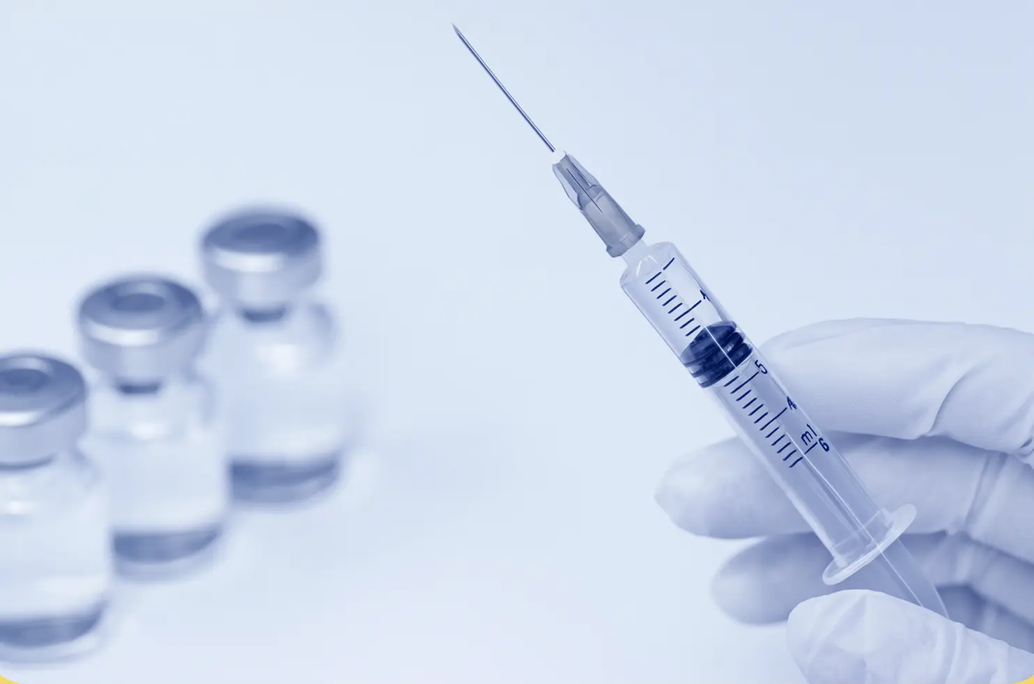 Gloved hand holding a syringe with vaccine vials blurred in the background.