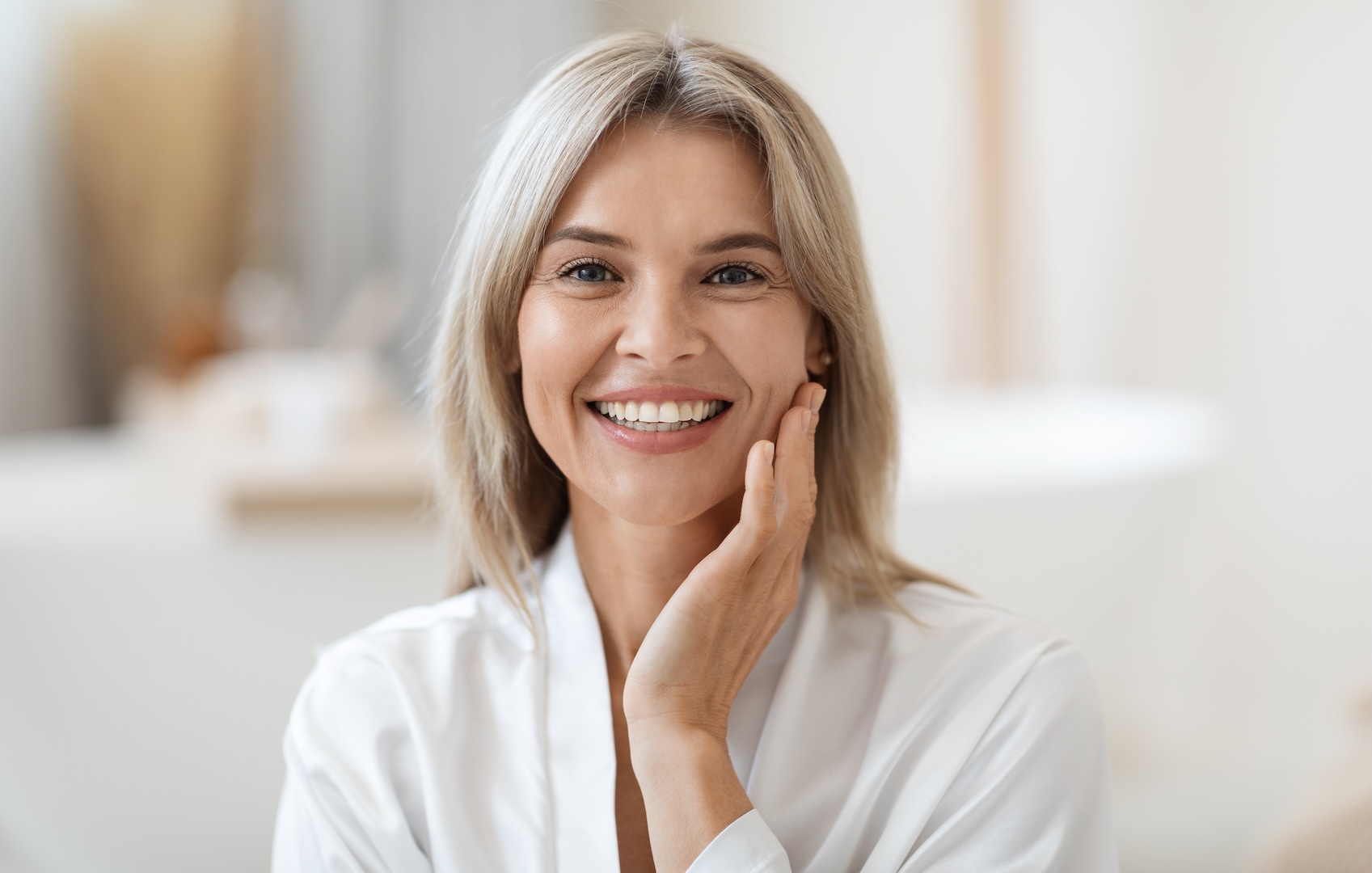 Smiling middle-aged woman with blonde hair touching her cheek in a bright room.