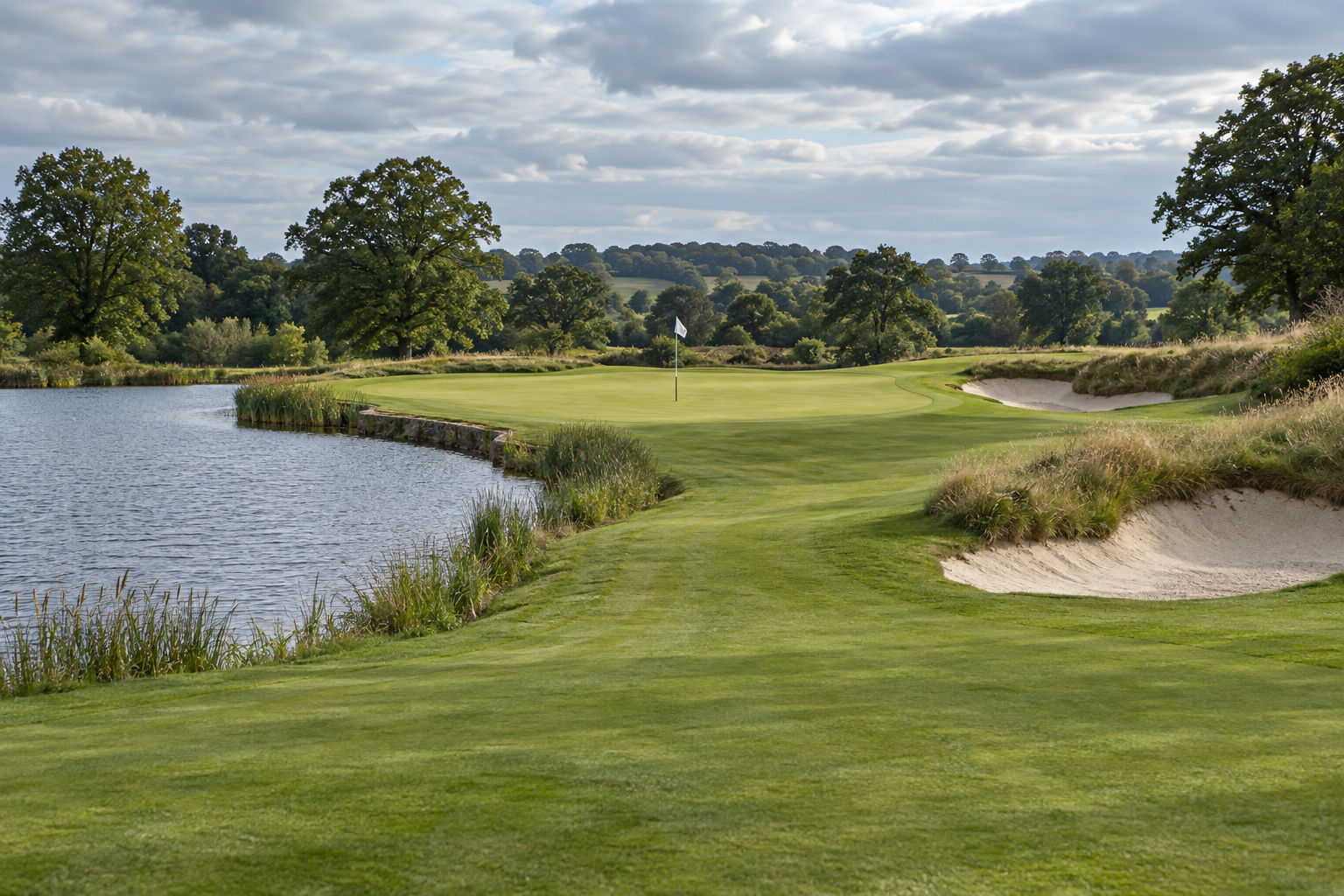 An approach to a golf green, with water on the left, bunkers on the right and a white flag ahead