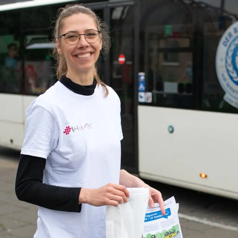 Lächelnde Frau mit Brille in einem weißen „Hürth“-T-Shirt, die vor einem Bus Papiere in der Hand hält.